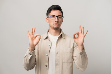 Unshakable confidence. A man in eyeglasses and a beige overshirt holding both hands in an OK gesture, exuding bold self-assurance. Modern style meets unstoppable charisma