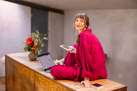 A fashionable woman in a red outfit sits on a wooden countertop with a laptop and smartphone, embracing a modern work-from-home lifestyle. Warm lighting and fresh flowers add charm to the cozy setting
