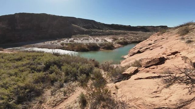 Virgin River Gorge- Hurricane, Utah, USA - February 10, 2025: FPV-First Person View-Descending over a rocky brushy hillside to a shallow river with sunlight glinting off the rapids 