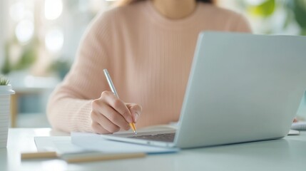 Person casually writing notes, young woman in light sweater focused on laptop, bright workspace with greenery behind.