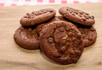 A group of warm chocolate cookies is displayed on a wooden surface, featuring a rich texture and dotted with chocolate chips, surrounded by a classic red and white checkered backdrop