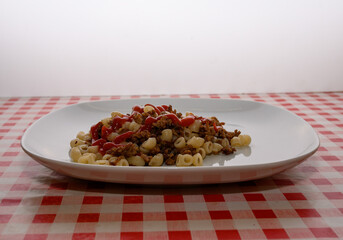 A plate features a hearty serving of cooked pasta mixed with seasoned meat and topped generously with ketchup, resting on a classic red and white checkered tablecloth