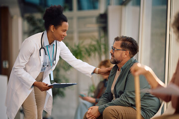 Happy African American female doctor talking to mature man in waiting room at medical clinic.