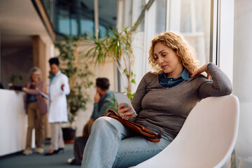 Smiling woman using cell phone while waiting for doctor's appointment at clinic.