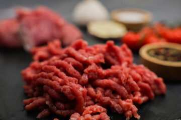 Close-up of raw red ground beef minced in a meat grinder lying on a black board, surrounded by out-of-focus cut meat, vegetables and seasonings in wooden bowls. The concept of diet, proper nutrition