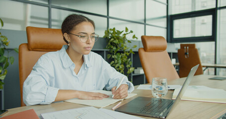 A young professional woman writing notes at a modern office desk with laptop, documents, and stationery in a bright workspace with large windows and contemporary furniture