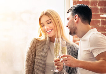 A young couple enjoys a joyful moment together, toasting with champagne glasses while sharing smiles in a warm, inviting indoor atmosphere filled with natural light.