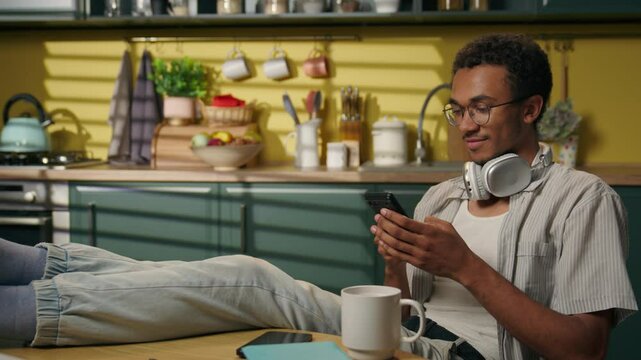 African American man ethnic guy male freelancer businessman gen z teen student studying at home kitchen browsing mobile phone internet social media scrolling message online shopping app order delivery