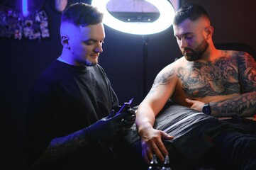 bearded tattoo artist working at his studio tattooing sleeve on the arm of his male client. Man getting tattooed by professional tattooist