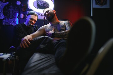 Male tattoo artist holding a tattoo gun, showing a process of making tattoos on a male tattooed model's arm