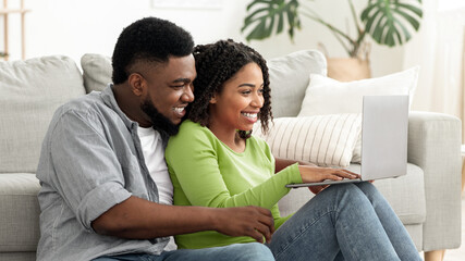 Fototapeta premium A happy black couple sits on a couch, focused on their laptop. They share smiles and laughter, enjoying their time together in a bright, comfortable living space with plants around.