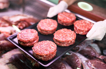 Close-up view of a tray with row burger patties held in hands by seller against background of showcase of butcher shop