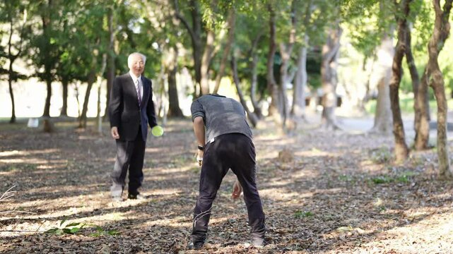 A winter park in Japan. A 78-year-old Japanese man in a suit stands in a natural park filled with trees, playing with his son in his 30s throwing colorful balls.