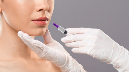 Obraz premium Aesthetic modern medicine concept. Lip injection for young woman over grey studio background, closeup. Unrecognizable cosmetitian injecting lip filler for beautiful lady looking at copy space