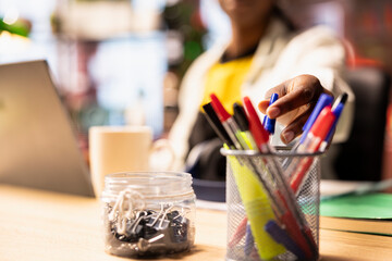 Close up of student in home office reaching for pen from desk organizer, preparing to jot down important notes. Focus on workspace stationary holder used by person in blurry background to do writing