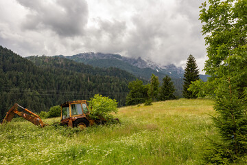 vista panoramica su un ambiente naturale di montagna nell'Italia settentrionale, di giorno, in estate, con cielo nuvoloso e coperto e con una vecchia ruspa abbandonata in un campo con erba alta