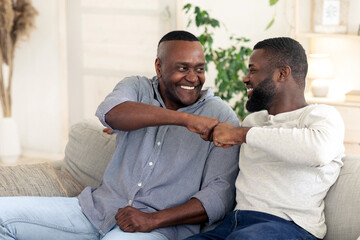 Two men are sitting on a couch in a bright room, laughing and sharing a fist bump. They express joy and camaraderie, showcasing a strong bond during a relaxed moment together.