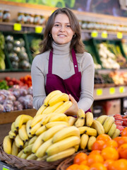 Young woman seller in apron lays out bananas on counter in vegetable shop