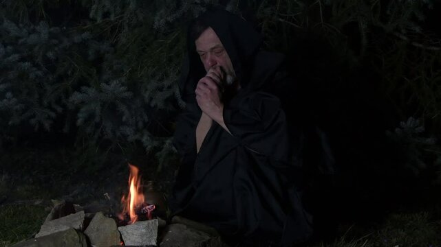 a mysterious hooded monk prays by the fire with a