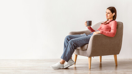 A young woman with brunette hair sits in a cozy armchair, holding a cup. The room has a simple,...