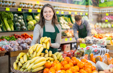Young positive female seller in apron displaying assortment of bananas at supermarket