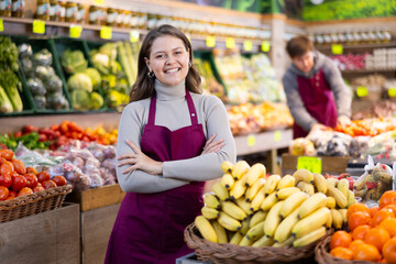 Young positive female seller in apron displaying assortment of bananas at supermarket