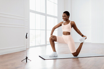 A young African American woman performs a stretching exercise on a yoga mat in her bright home. She...