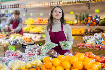 Young woman seller in apron lays out green peas on counter in vegetable shop