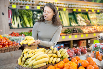 Young woman shopper in casual clothes chooses bananas in vegetable shop