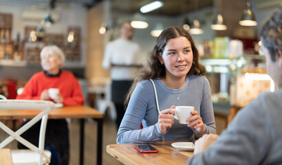 Fototapeta premium Girl in cafe sit opposite male colleague in cafe, they chat and joke, drinking coffee, spend time. Date, romantic meeting, nice time together. Atmosphere of cafe, restaurant conducive communication