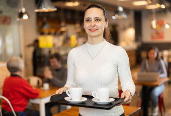 Woman visitor in self-service cafeteria carries tray with two cups of coffee. Customer has purchased freshly brewed coffee for herself and companion