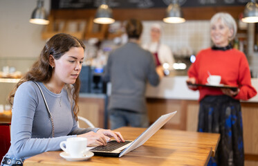 During lunch break, girl spends time with laptop at table in cafe, drinks coffee, and communicates via video link with relatives.