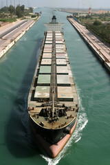 A cargo ship is seen traveling through a narrow canal, surrounded by calm water and green landscapes. The serene setting highlights maritime transportation and the importance of shipping routes.