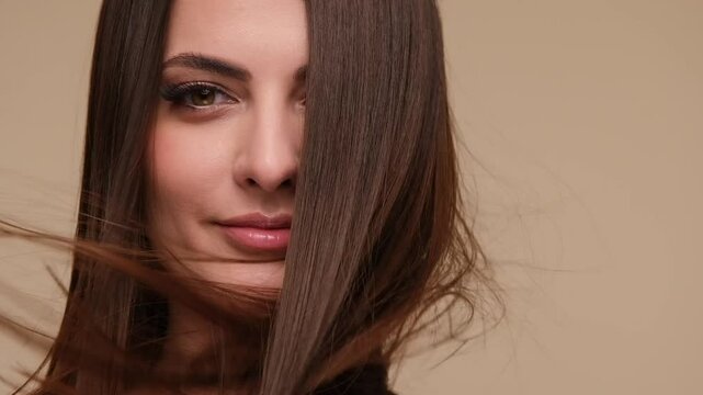 Beautiful woman with brown healthy hair on beige background. Wind blows on beauty hair.
