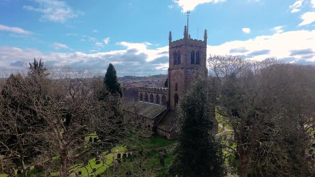 Aerial drone shot of Holy Trinity Church of England Church in Eccleshall, Staffordshire, UK on a spring afternoon. The sun is shining as we pan past the graveyard and cricket club. 