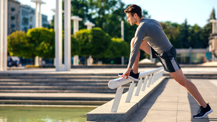 Fototapeta premium Morning stretching. Athletic guy in sportswear puts his foot on railing and does exercises in park, side view, free space