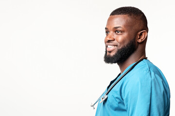 A confident African American man doctor in blue scrubs smiles while standing in a clinic. His stethoscope is draped around his neck, showcasing his commitment to patient care and health, copy space