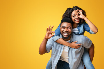 Fototapeta premium A joyful couple smiles and poses enthusiastically, with the woman riding on the man's back. The bright yellow backdrop adds a cheerful ambiance to their fun interaction.