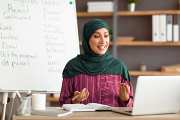 A young Middle Eastern teacher in hijab engages with students in an online English lesson. She appears enthusiastic and knowledgeable, with educational materials visible in the background.