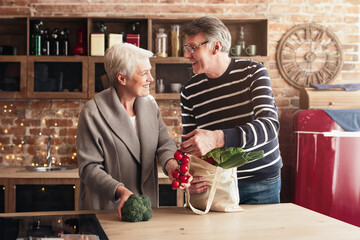 Cheerful senior couple unpacking shopping bags with food in kitchen, have grocery together, free space