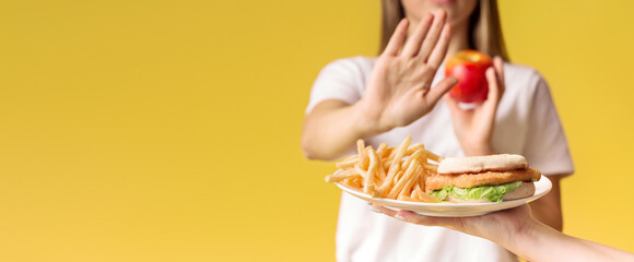 A person stands against a vibrant yellow background, refusing a plate of fries and a sandwich,...