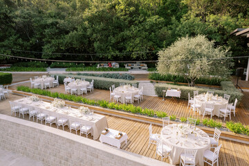 Festive tables with bouquets of flowers stand on a wooden deck in the garden behind a stone fence. Drone