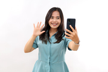 Fototapeta premium Smiling pleasant looking Asian girl waves palm and says hello to camera of modern smartphone, makes video call, has long dark hair, wears casual shirt, poses indoor.