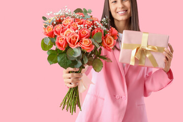 Young woman holding bouquet of beautiful flowers and gift box on pink background