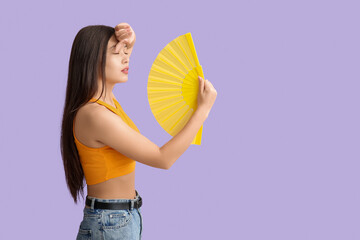 Young woman with hand fan suffering from heat on lilac background