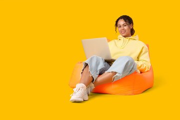 Young African-American QA engineer with laptop sitting on beanbag chair against yellow background