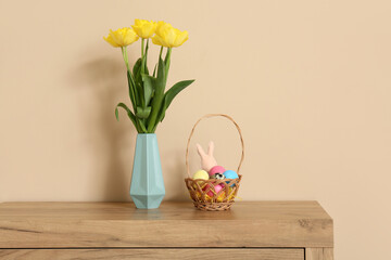 Tulip flowers in vase with wicker basket of colorful eggs and paper rabbit on commode near beige...
