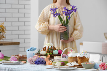 Woman holding vase with flowers near table setting for Easter celebration in kitchen. Closeup