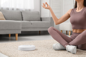 Sporty young woman meditating with robot vacuum cleaner on carpet at home