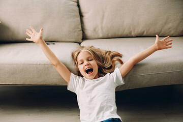 Joyful little girl sitting on the floor with arms raised high, couch in background, expressing excitement indoors
